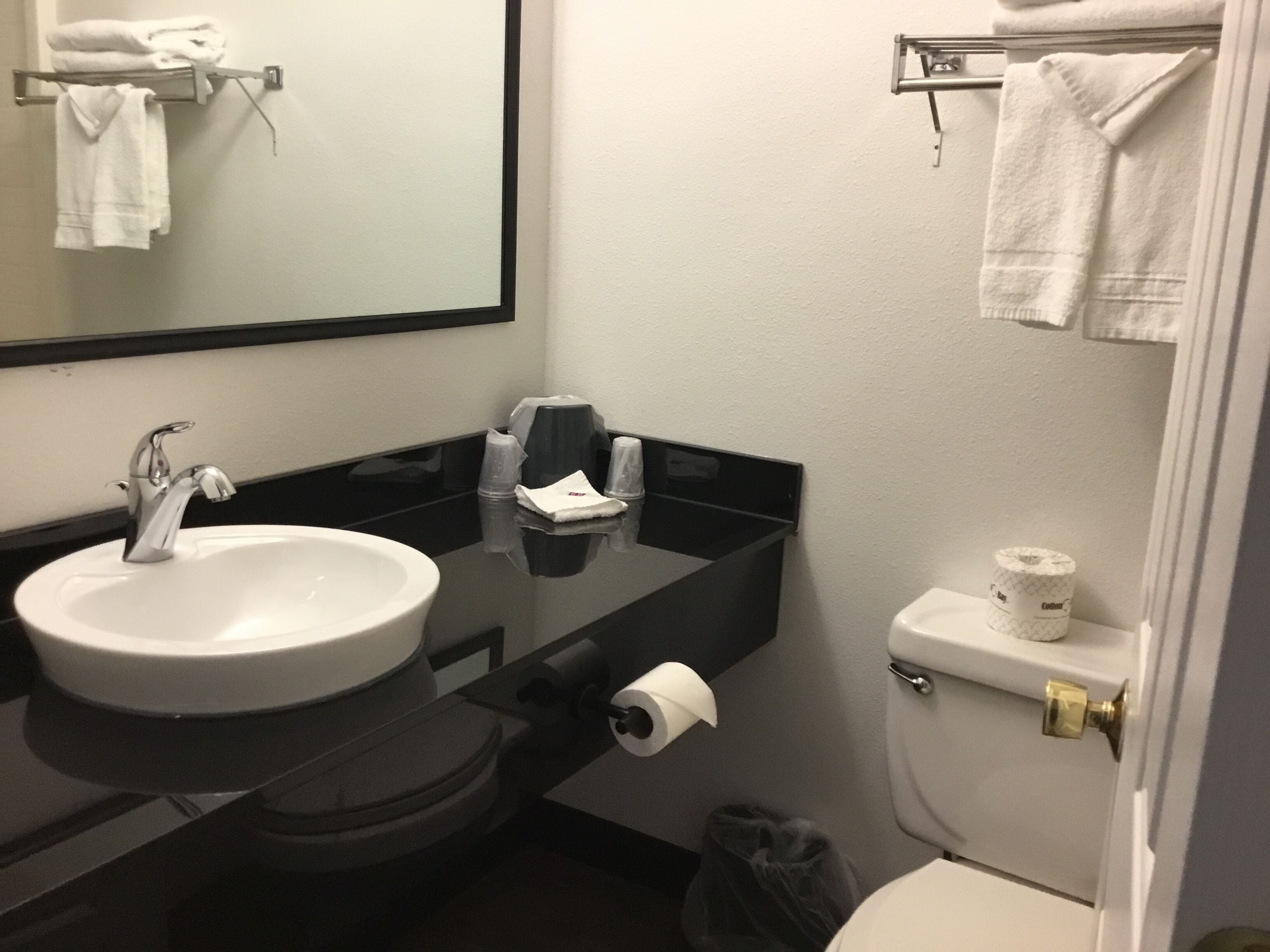 A bathroom with a white sink, black countertop, and a toilet. There are towels on a rack and a trash can visible.