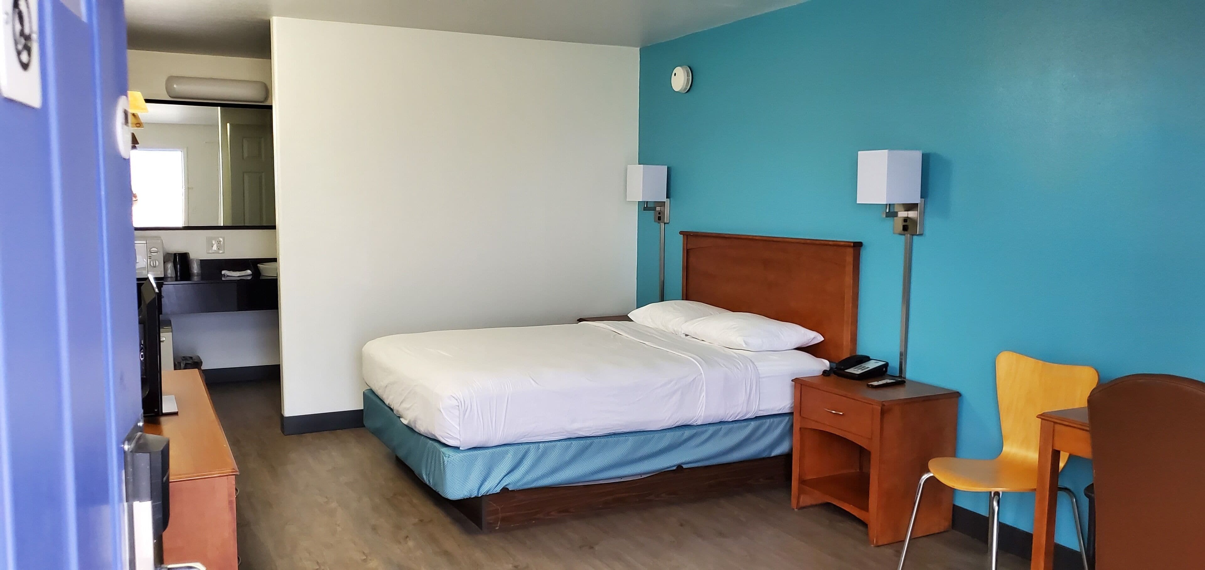 A hotel room with a bed, bedside table, chair, and a desk with a lamp.