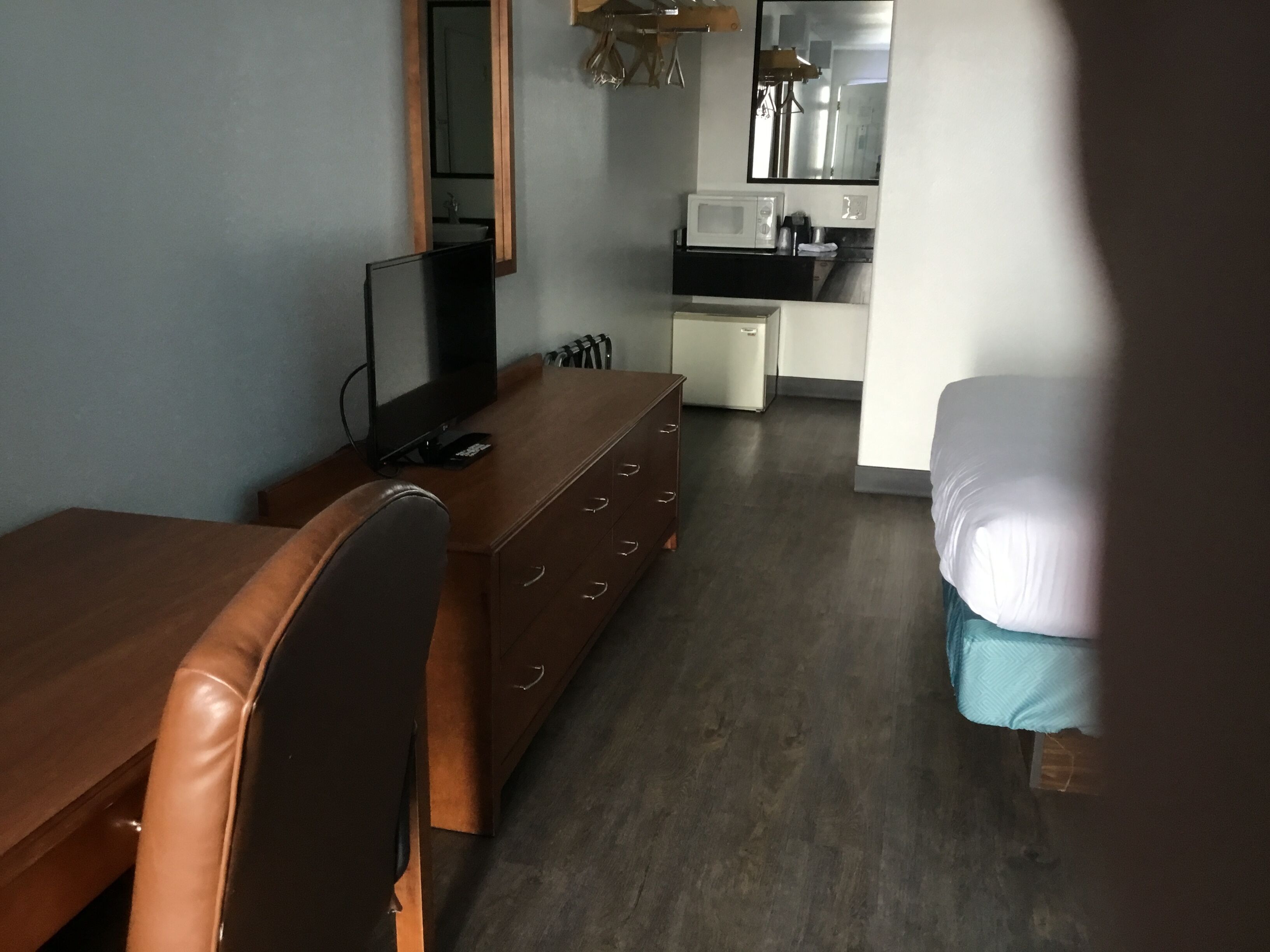 A compact hotel room with a wooden desk, a chair, a bed, a small kitchenette, and a mirror.