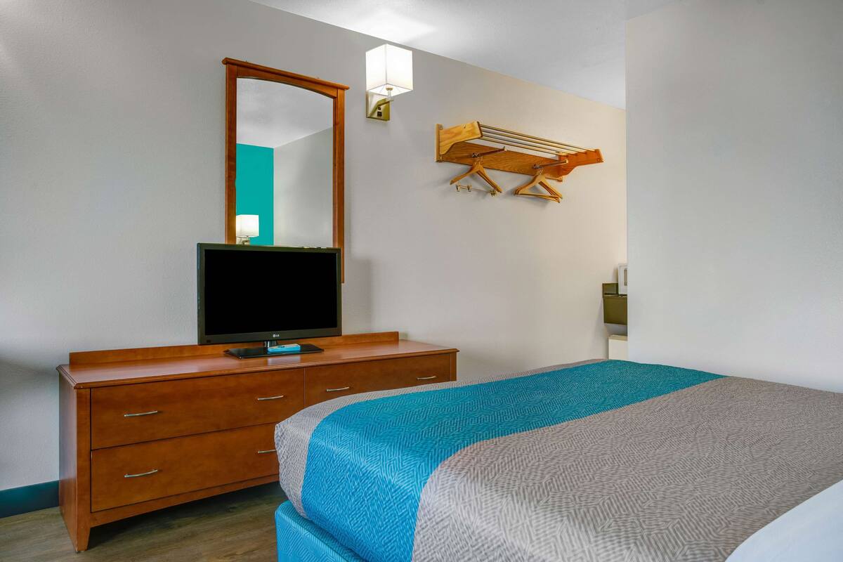 A hotel room with a wooden dresser, a flat-screen TV, a bed with patterned bedding, a wall-mounted mirror, and a wall-mounted drying rack.