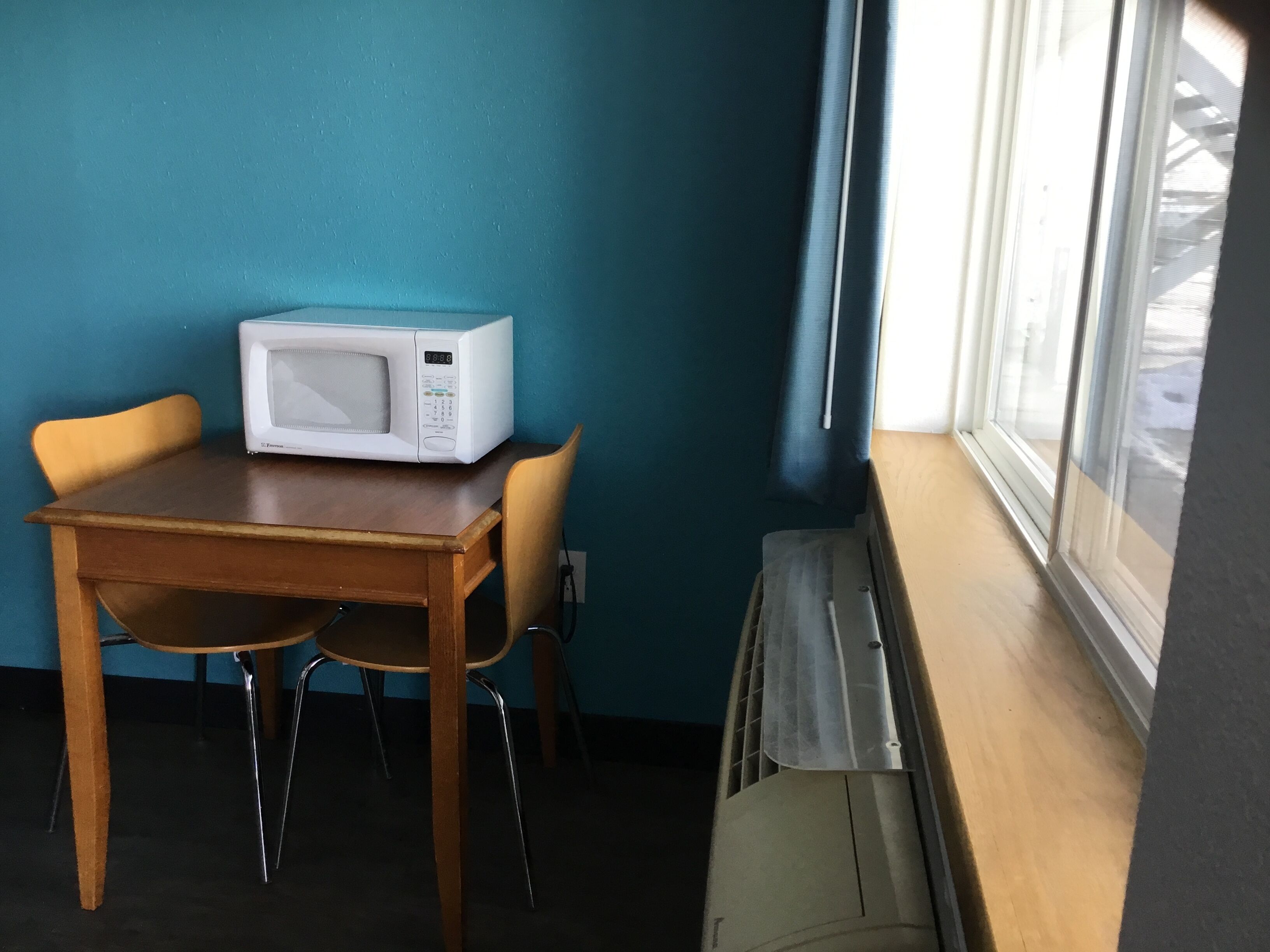 Una habitación con una mesa de madera, dos sillas, un microondas blanco y una ventana con persianas.