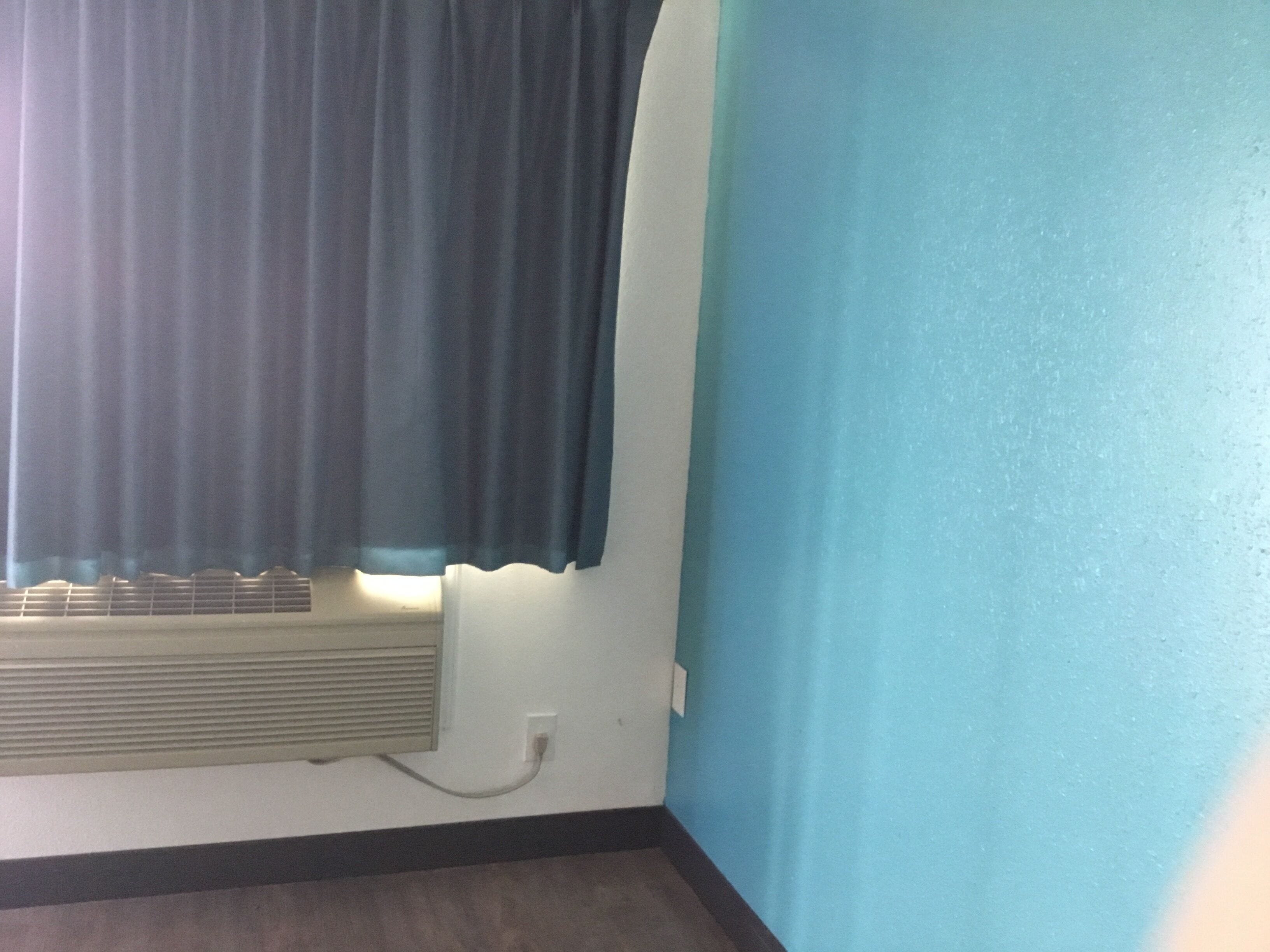 Una habitación con una ventana cubierta por cortinas oscuras, un aire acondicionado instalado en la pared y una pared azul.