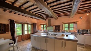 A spacious kitchen with a central island, wooden beams on the ceiling, and large windows providing natural light. - Col Di Forche (Monterchi)