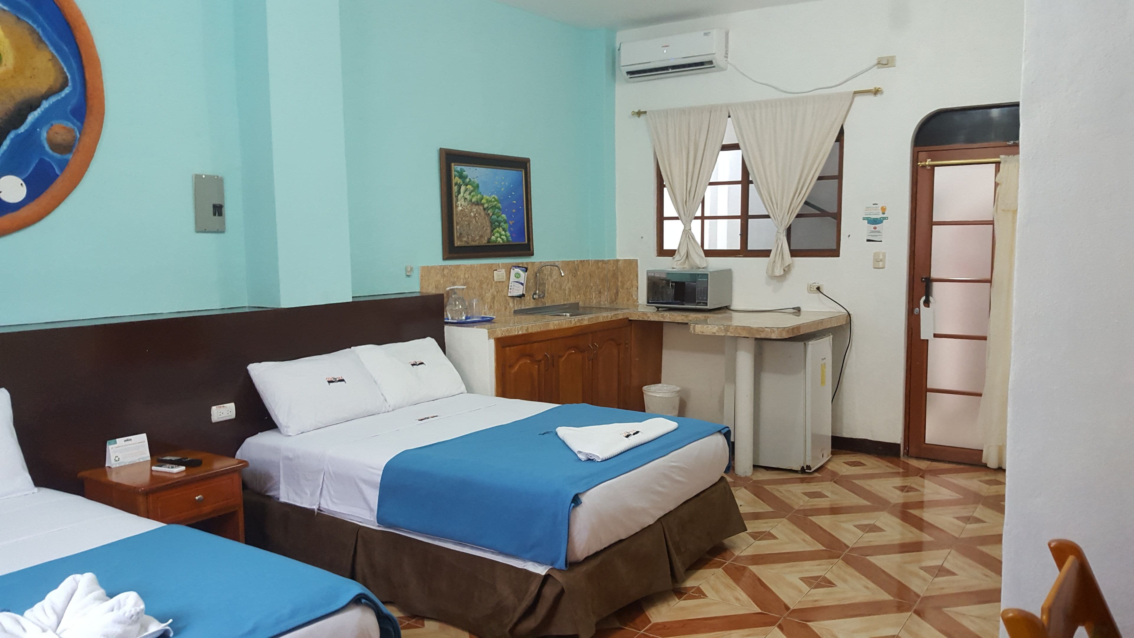 A hotel room with two beds, a small kitchenette, a TV, and a window with curtains.