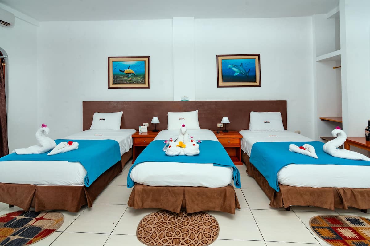 A hotel room with three beds, each with a blue bedspread and brown headboard. There are two framed pictures on the wall, a bottle of wine, and a glass on the nightstand.