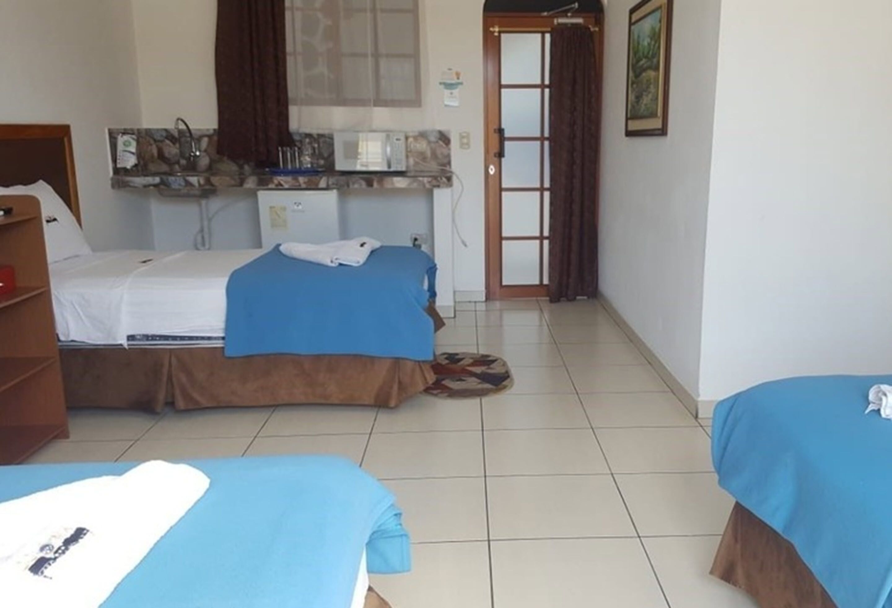 A hotel room with two beds, a small kitchenette, a bathroom with a shower, and a door leading to another room.