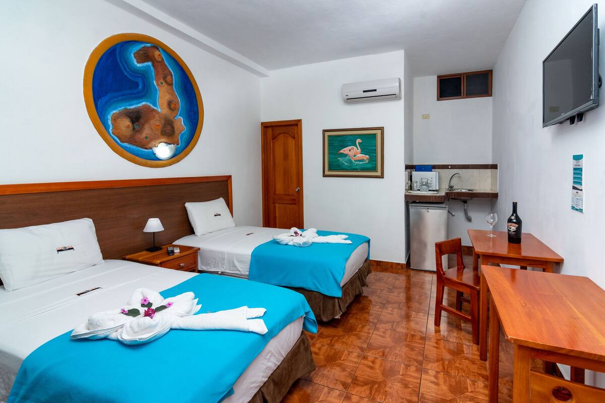 A hotel room with two beds, a wooden desk, a small kitchenette, and a TV mounted on the wall.