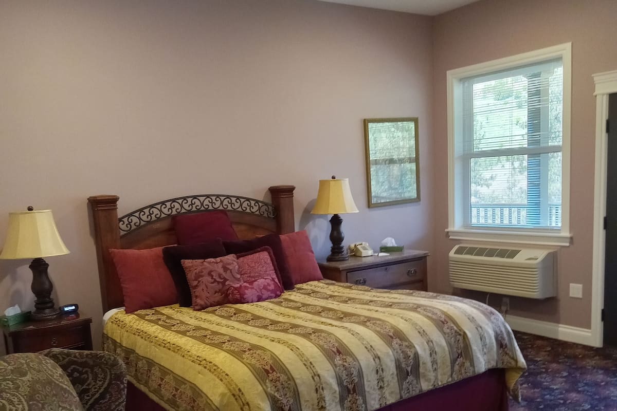 A bedroom with a large bed, two bedside lamps, a nightstand, a chair, a window with blinds, and a wall-mounted air conditioner.