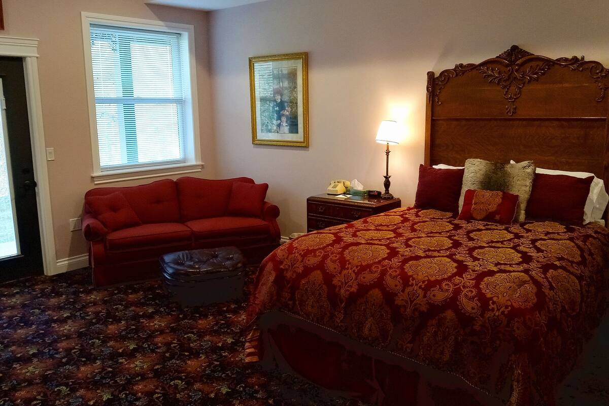 A bedroom with a large bed, a red sofa, a small table with a lamp, and a window with blinds.