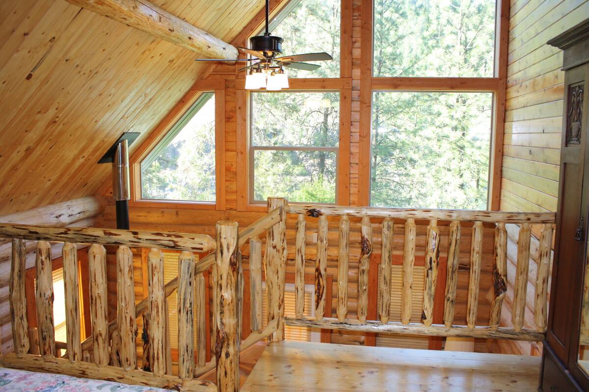 A wooden cabin with a loft bedroom, large windows, and a ceiling fan.