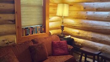 L’intérieur d’un chalet confortable, avec un plancher en bois, un canapé moelleux recouvert d’un tissu à motifs, une petite table avec une lampe et une fenêtre équipée de stores.