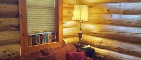 A cozy log cabin interior with a wooden floor, a cushioned sofa with patterned upholstery, a small table with a lamp, and a window with blinds.