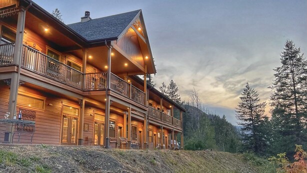 A large, multi-story wooden lodge with a balcony and outdoor seating area, surrounded by trees. - Hearthstone Elegant Lodge by the River (Kamiah)