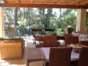 An outdoor patio with wicker furniture, a wooden table, and a view of a garden with trees and a fence. - Mariasole (Castiglione della Pescaia)