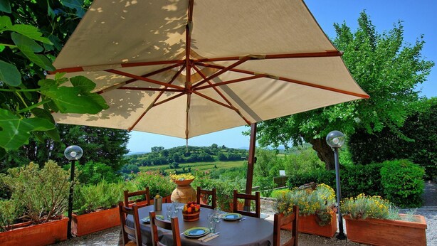 An outdoor dining area with a table set for four, surrounded by chairs, under a large umbrella. - Uccellaia (Magione)