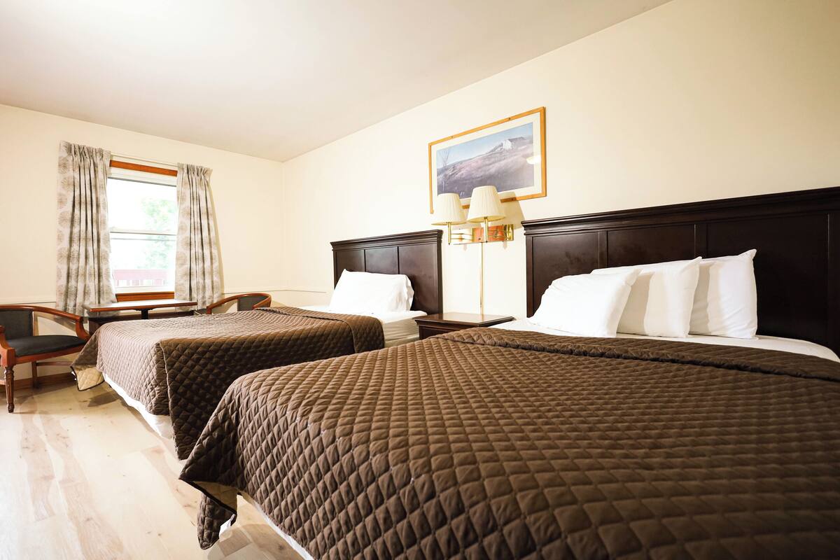 Ein Hotelzimmer mit zwei Queensize-Betten, einem Schreibtisch, einem Stuhl, einer Lampe und einem Fenster mit Vorhängen.