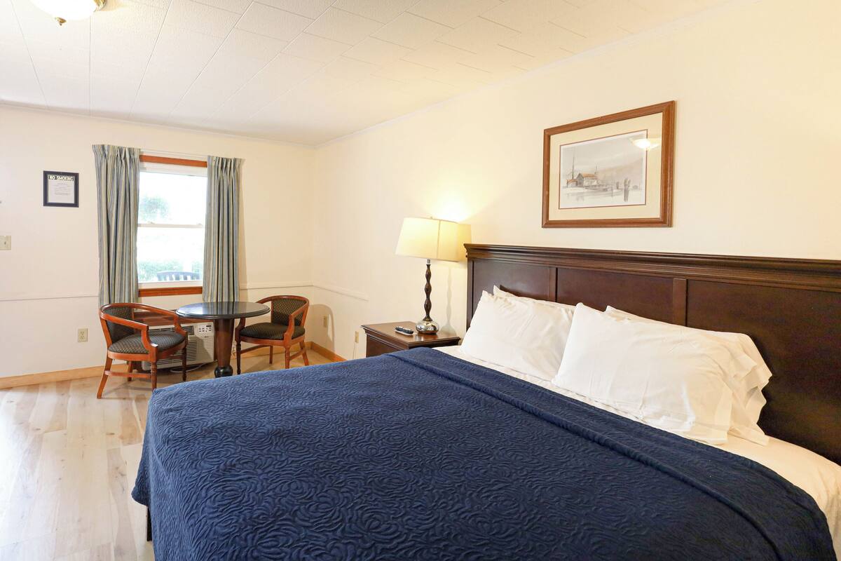 Ein Hotelzimmer mit einem großen Bett, einem Schreibtisch mit Stuhl, einer Lampe und einem gerahmten Bild an der Wand.