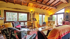 A cozy living room with wooden beams, comfortable seating, a television, and large windows offering a view of the outdoors. - Casa Vania (Reggello)
