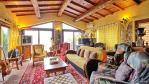 A cozy living room with a wooden ceiling, a stone fireplace, a large window, and a variety of seating including a sofa, chairs, and a coffee table. - Casa Vania (Reggello)