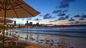 Une vue sur la plage, avec une ligne d’horizon urbaine en arrière-plan, une zone de sable mouillé et une chaise placée sous un parasol.