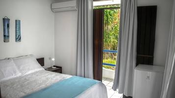 Een slaapkamer met een bed, nachtkastje, een luchtverkoeler aan de muur en uitzicht op een balkon met gordijnen.