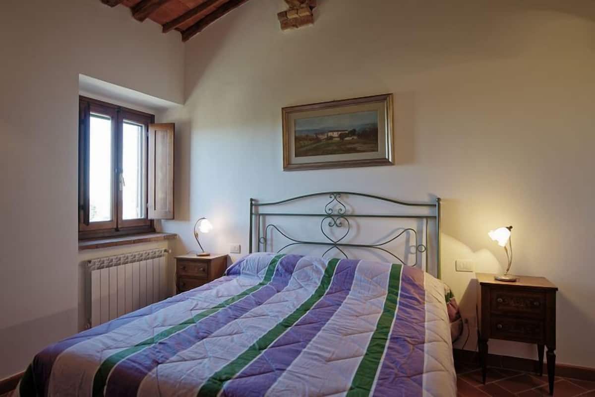 A bedroom with a bed, a window, a radiator, a nightstand, a lamp, and a painting on the wall.