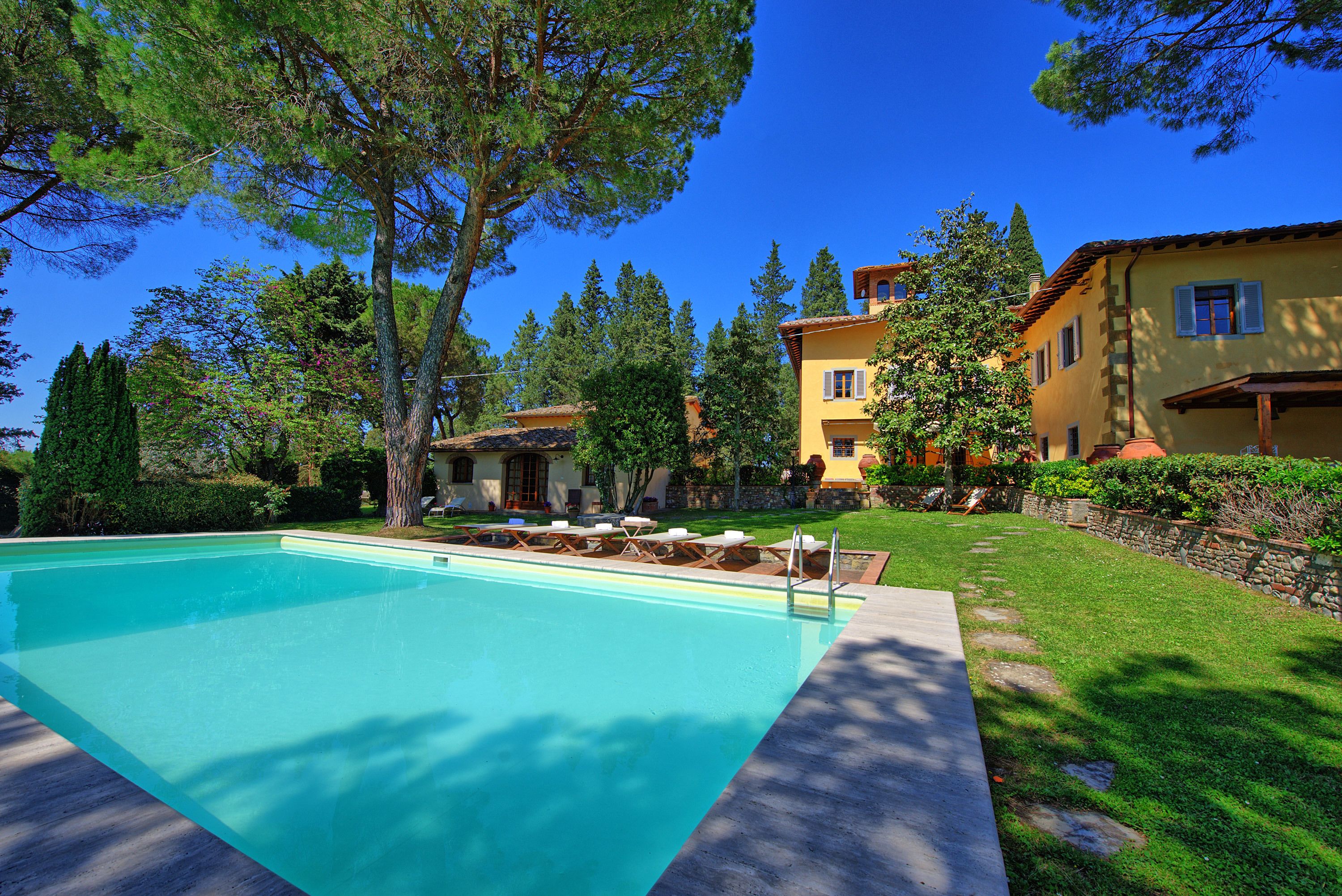 Una piscina amb gandules, envoltada per un jardí ben cuidat i arbres, amb un edifici groc al fons.