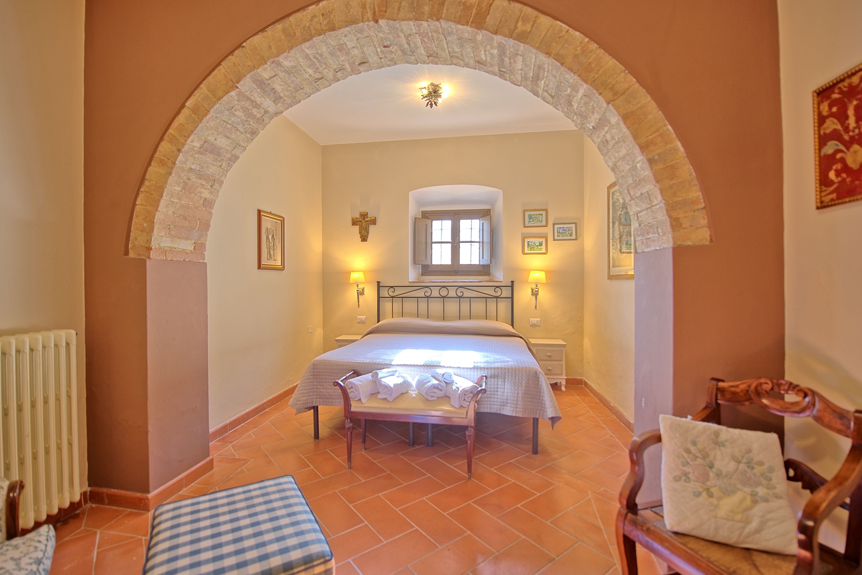A bedroom with a bed, a bench, a chair, a radiator, and a window.