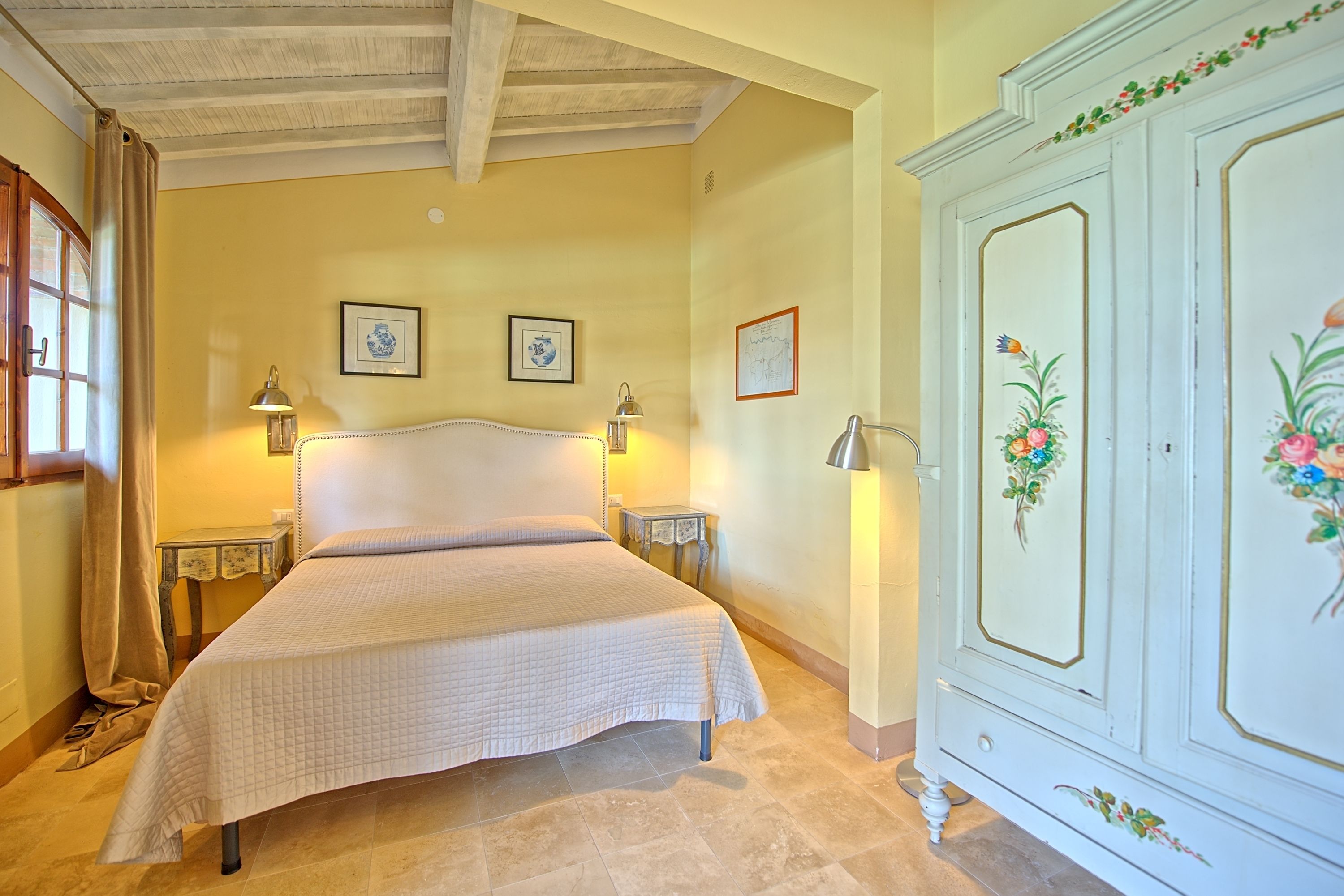 A bedroom with a large bed, two bedside tables with lamps, a wardrobe with floral decorations, and two framed pictures on the wall.
