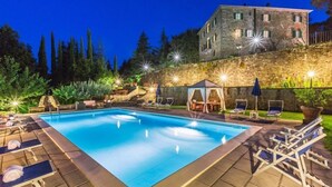 A rectangular swimming pool with lounge chairs and umbrellas, surrounded by a stone wall and a building with multiple windows. - Palazzo Rosadi (Monterchi)