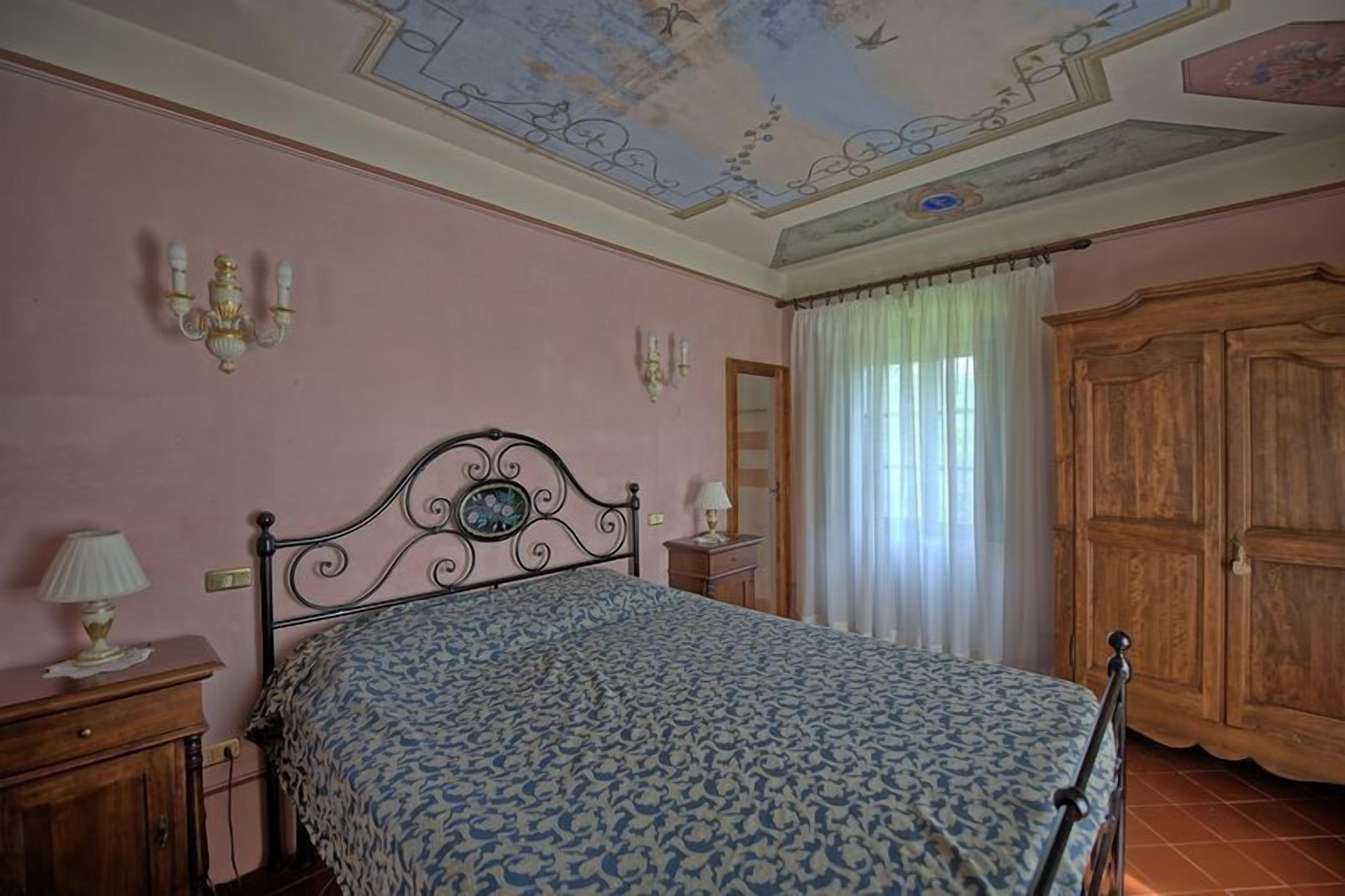 Une chambre à coucher comprenant un lit, une table de chevet, une armoire en bois et un plafond orné d’un motif peint.