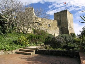 A historic stone building with a flag on top, surrounded by lush greenery and a paved area with steps leading up to it.