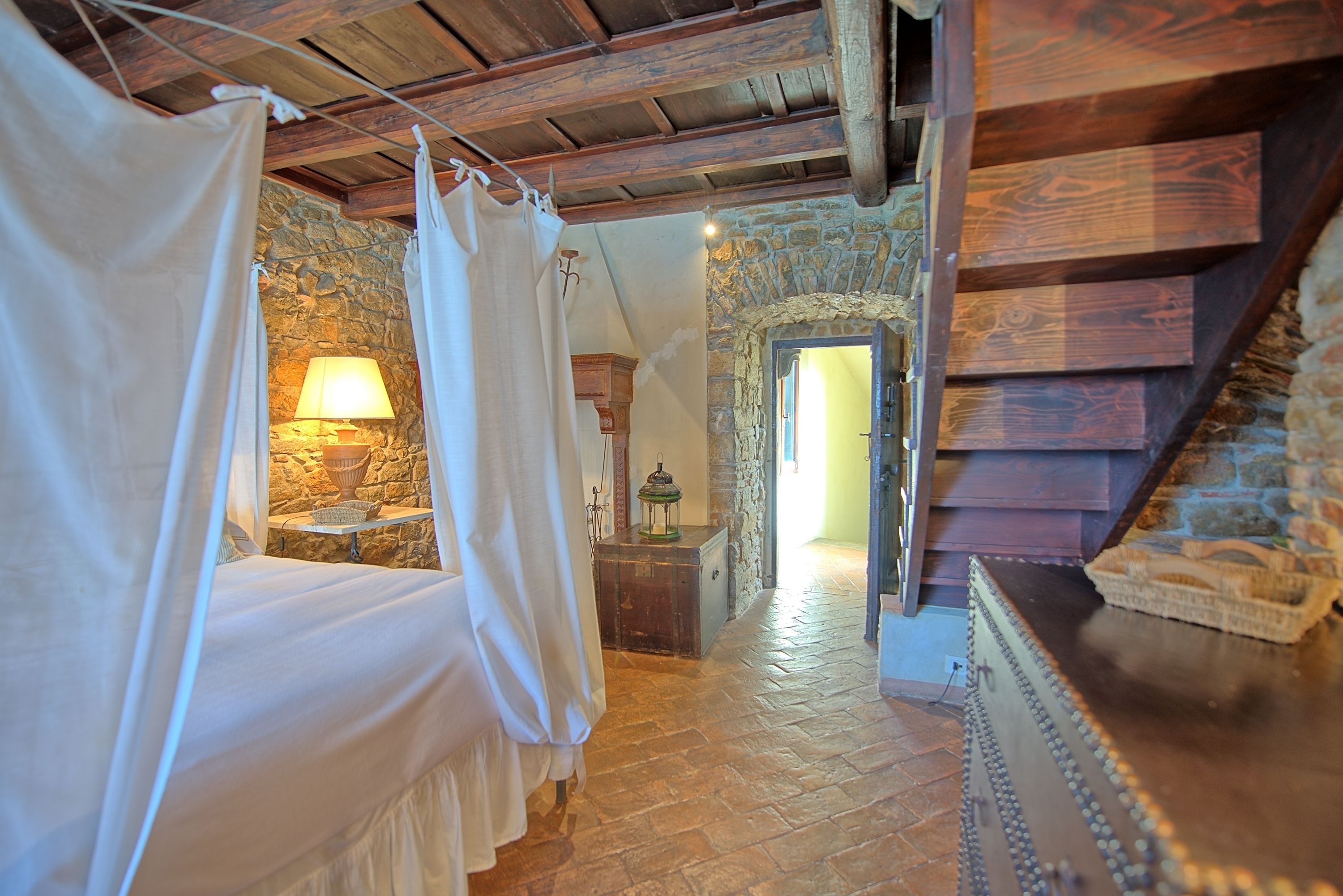 Un dormitorio rústico con pared de piedra, vigas de madera, cama con cortinas blancas, mesita de noche con lámpara y un cofre de madera.