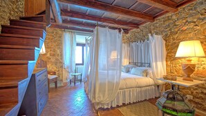 A bedroom with a stone wall, wooden staircase, a bed with a canopy, a bedside table with a lamp, and a small table with a basket. - Presidio (Orbetello)