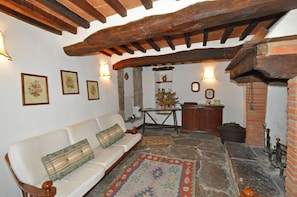 A rustic interior with exposed wooden beams, a white sofa with patterned cushions, a stone floor, and a traditional fireplace. - Borgo Del Castagno (Cortona)