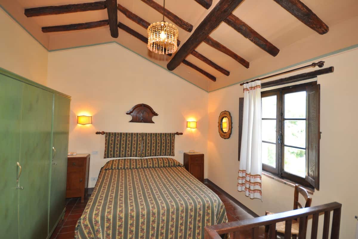 Un dormitorio con cama, vigas de madera, una araña, una ventana con cortinas y un armario de madera.