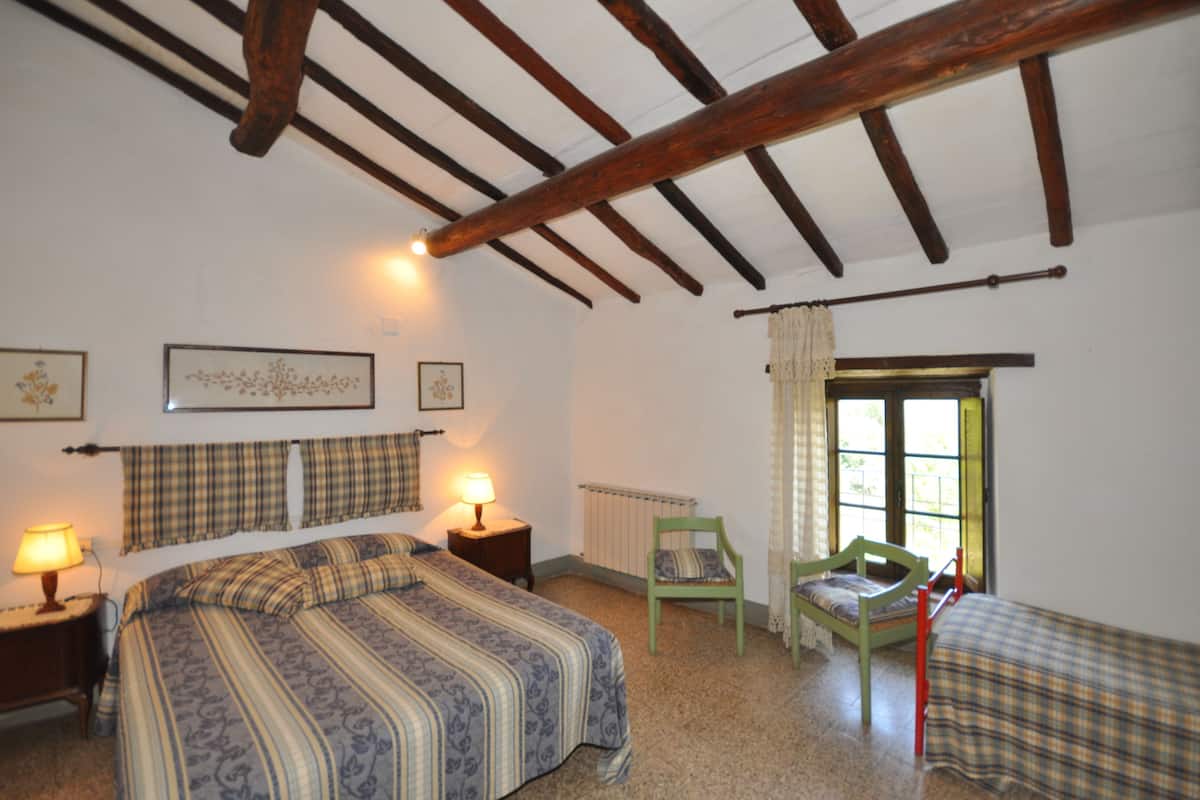 Un dormitorio con una cama, dos mesitas de noche, una silla, una ventana con cortinas y vigas de madera en el techo.
