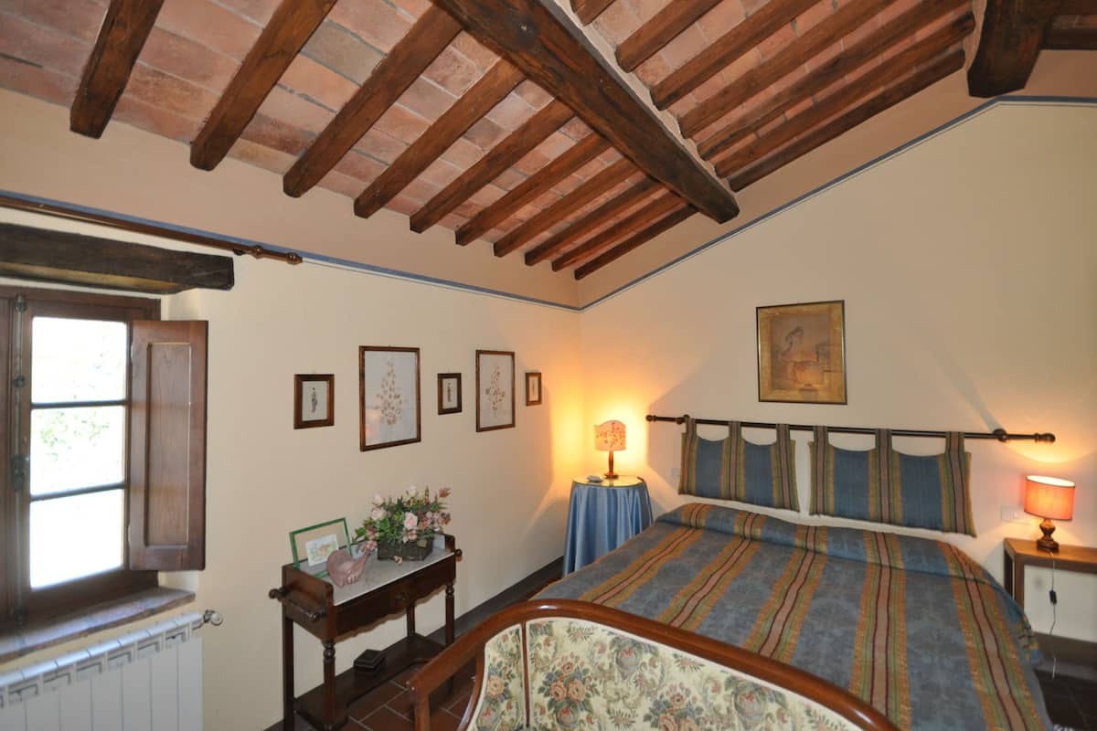Un dormitorio con techo de madera, una cama con ropa de cama a rayas, una mesita de noche con una lámpara y una ventana con vistas al verdor.