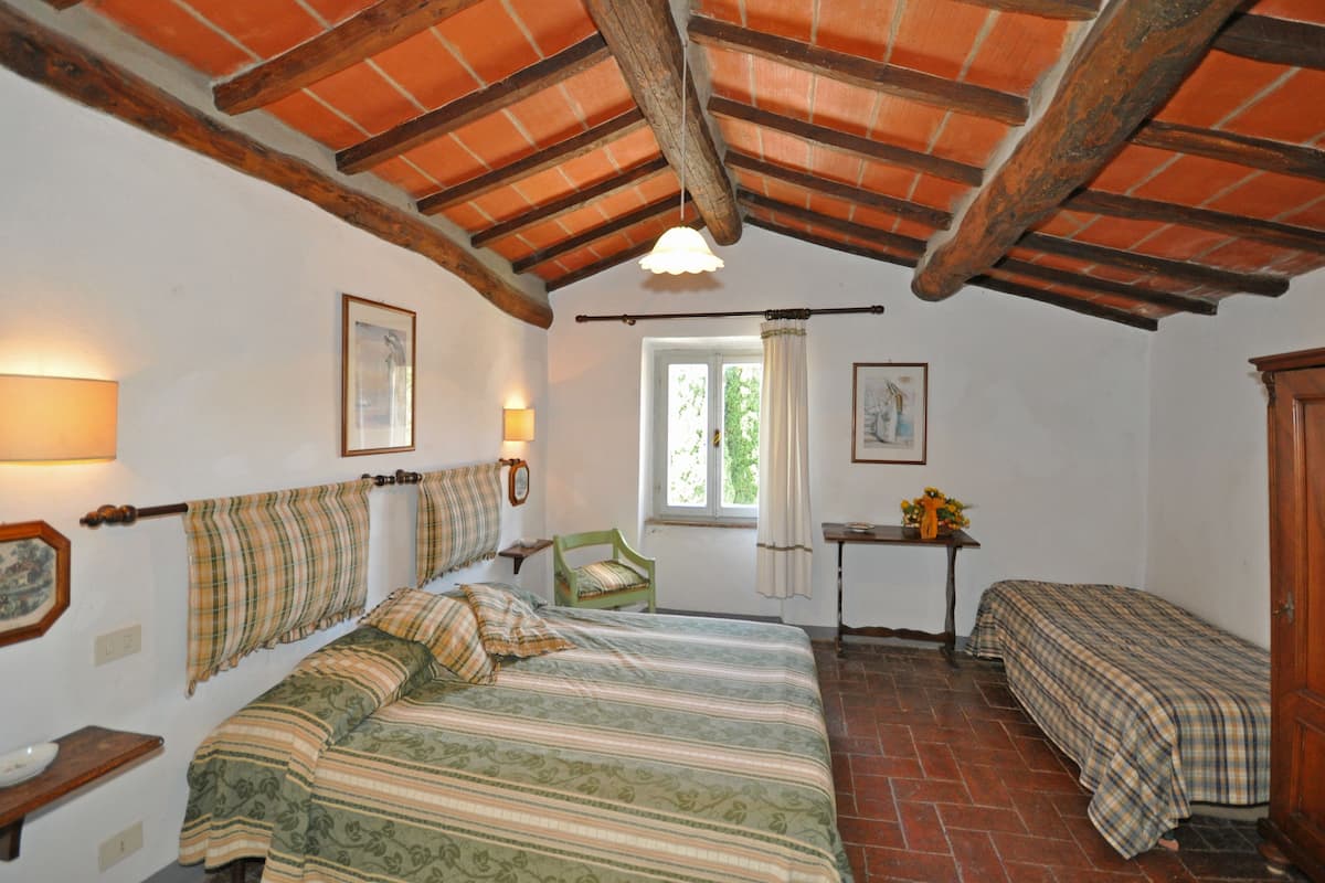 Un dormitorio con una cama grande, una cama más pequeña, una ventana con cortinas, techo de madera y una mesita con un jarrón.