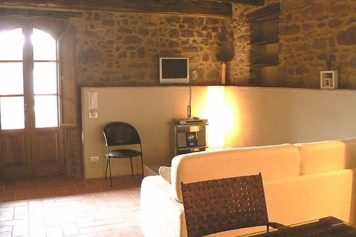 Una habitación rústica con pared de piedra, puerta de madera, televisor montado en la pared, una mesita con lámpara y un sofá blanco.
