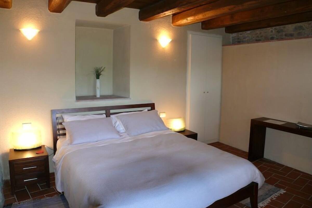 Un dormitorio con techo de madera, una cama con ropa de cama blanca, dos mesitas de noche con lámparas y una pequeña planta en una mesita de noche.