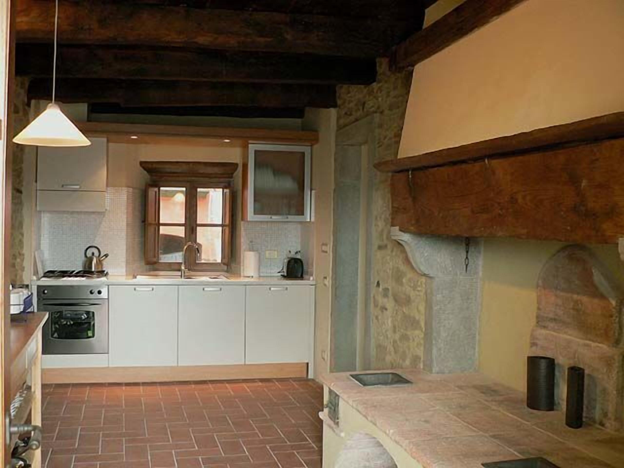 Cocina compacta con piso de ladrillo, armarios blancos, una lámpara colgante y una chimenea de piedra.