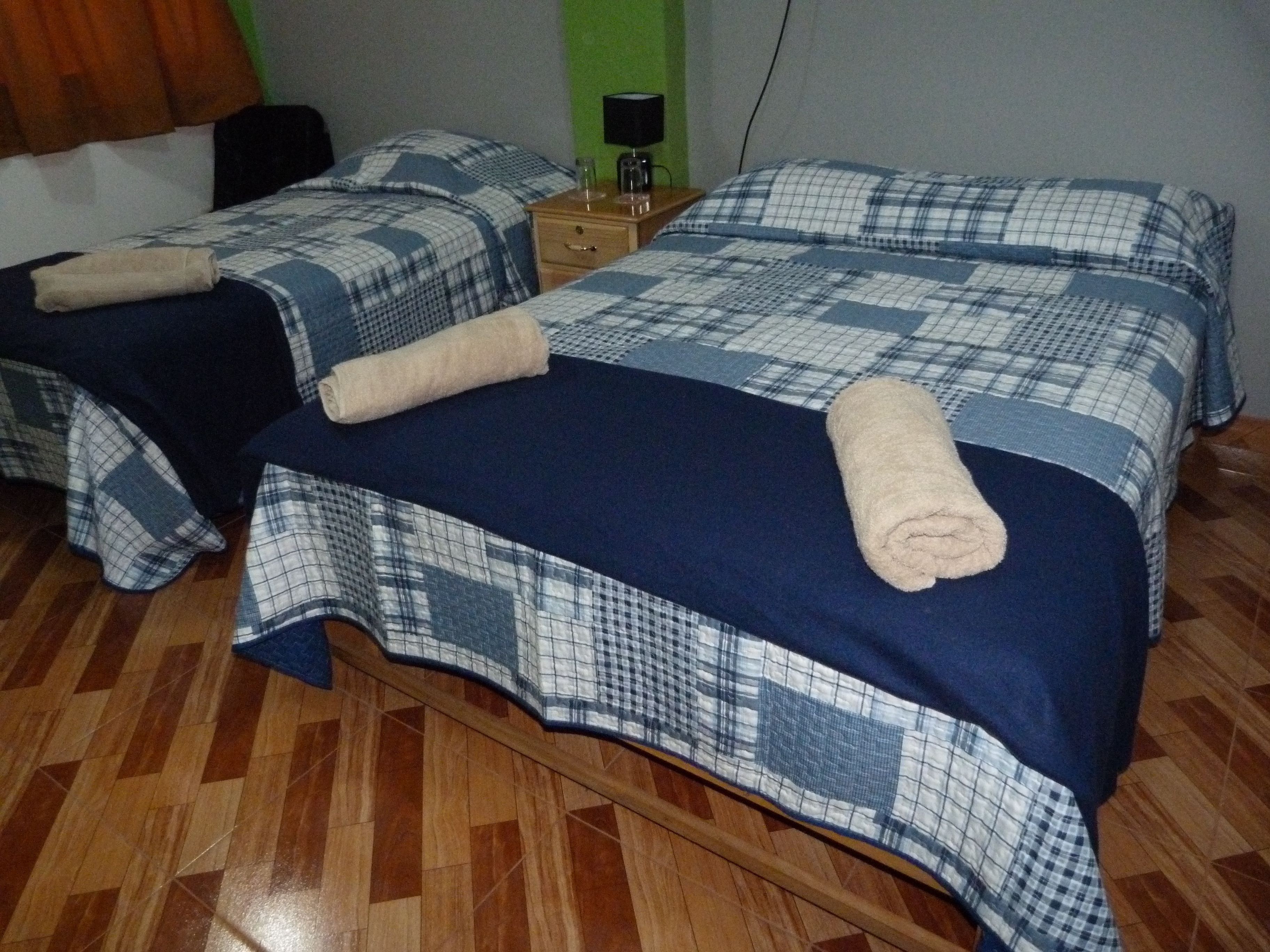 Una habitación con dos camas, cada una con una colcha a cuadros azules y blancos y una toalla enrollada. Hay una mesita de noche de madera con una lámpara entre las camas.