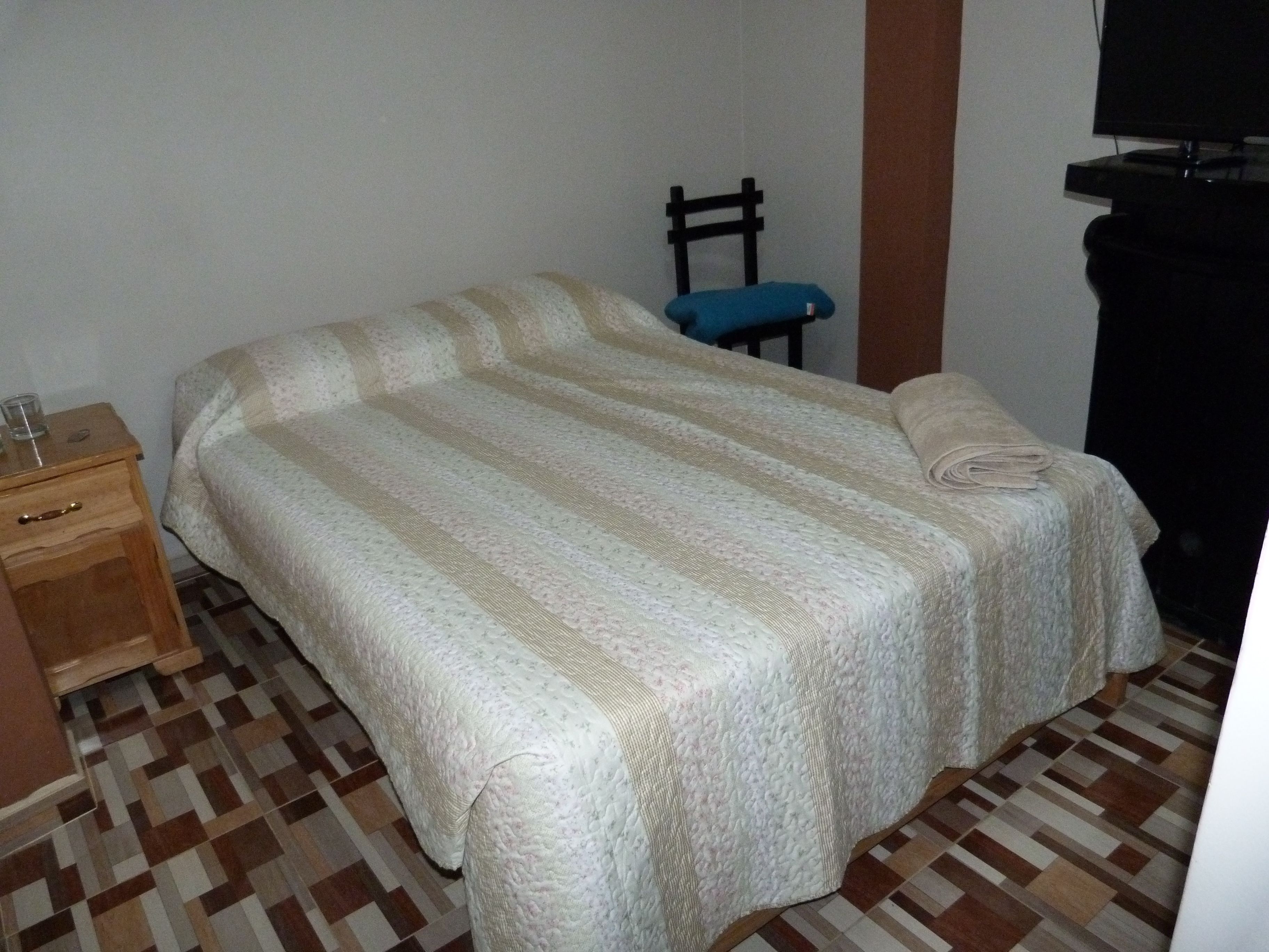 A neatly made bed with a striped bedspread, a wooden nightstand with a glass and a small item on it, a blue chair, and a television on a stand.