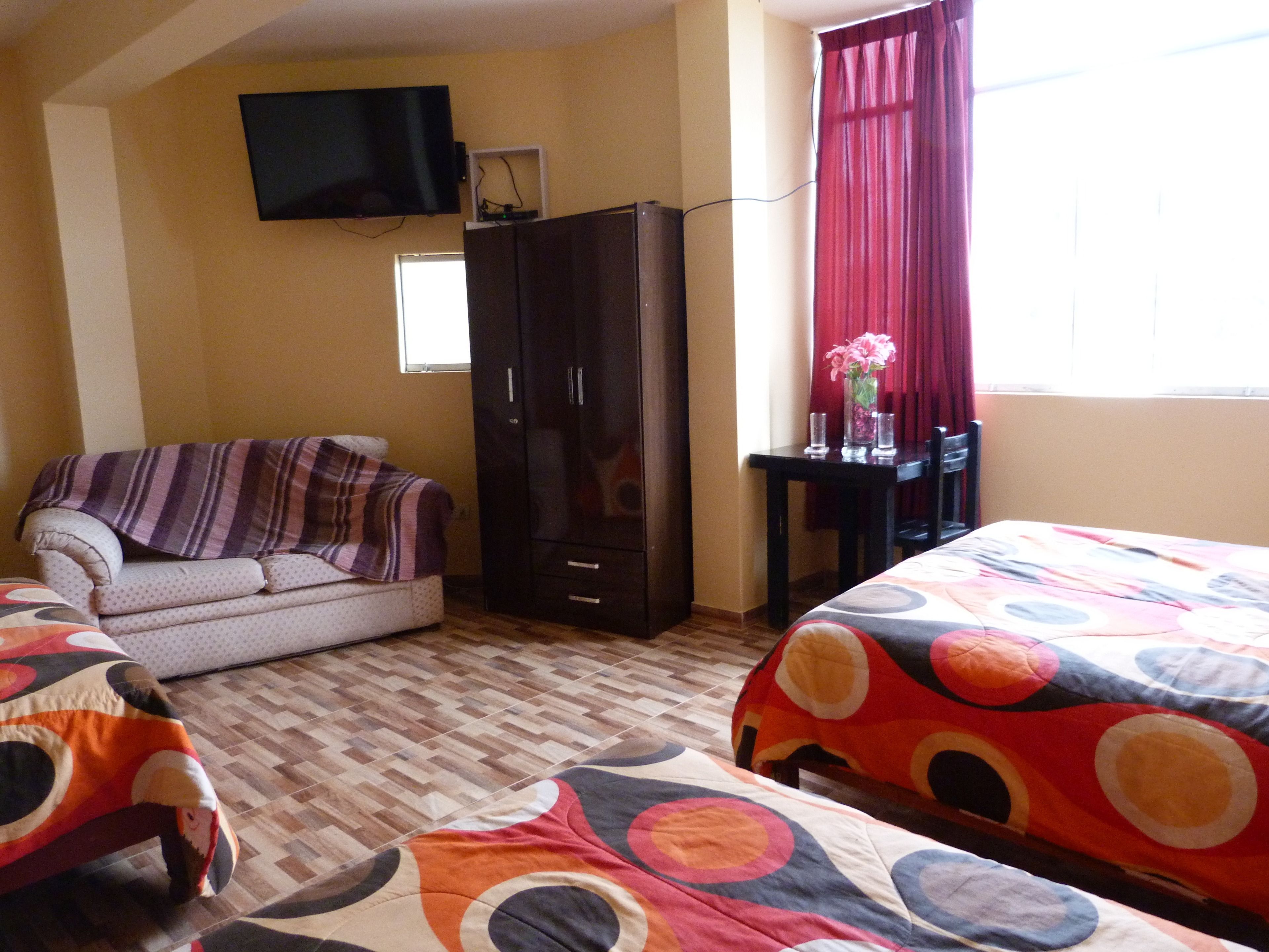 Un dormitorio con sofá, cama con edredón colorido, armario, escritorio con silla y televisión instalada en la pared.