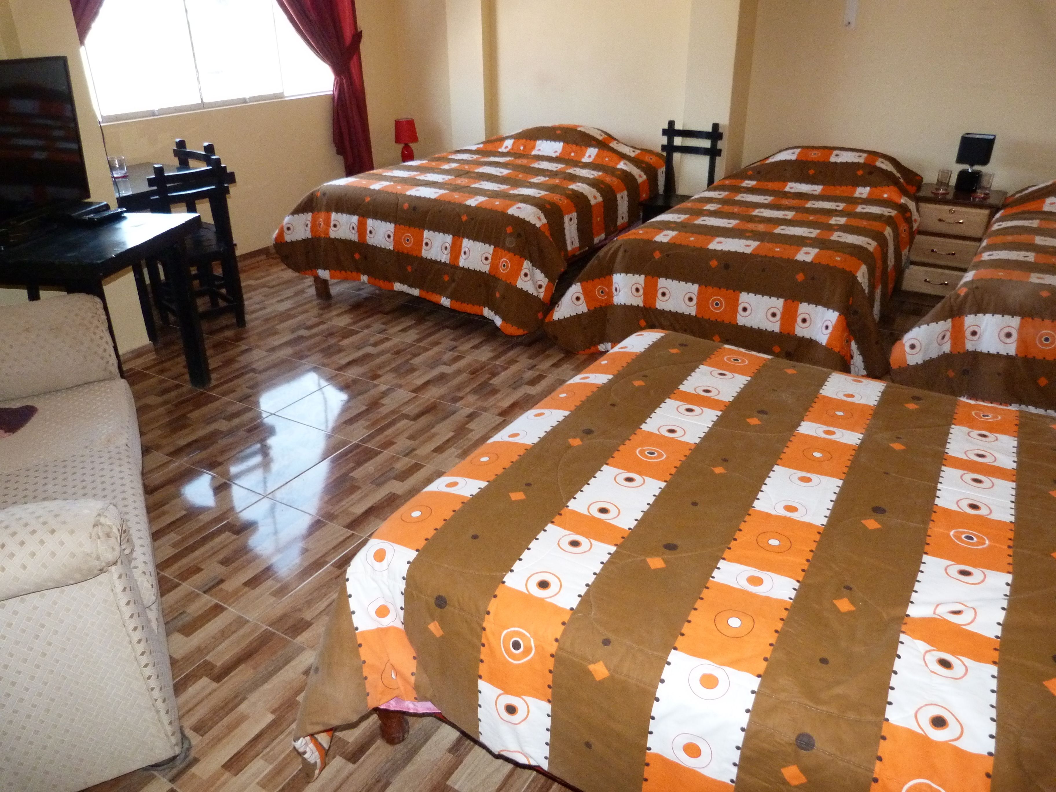 Quarto de hotel com três camas, piso de madeira, escrivaninha com cadeira e janela com cortinas.