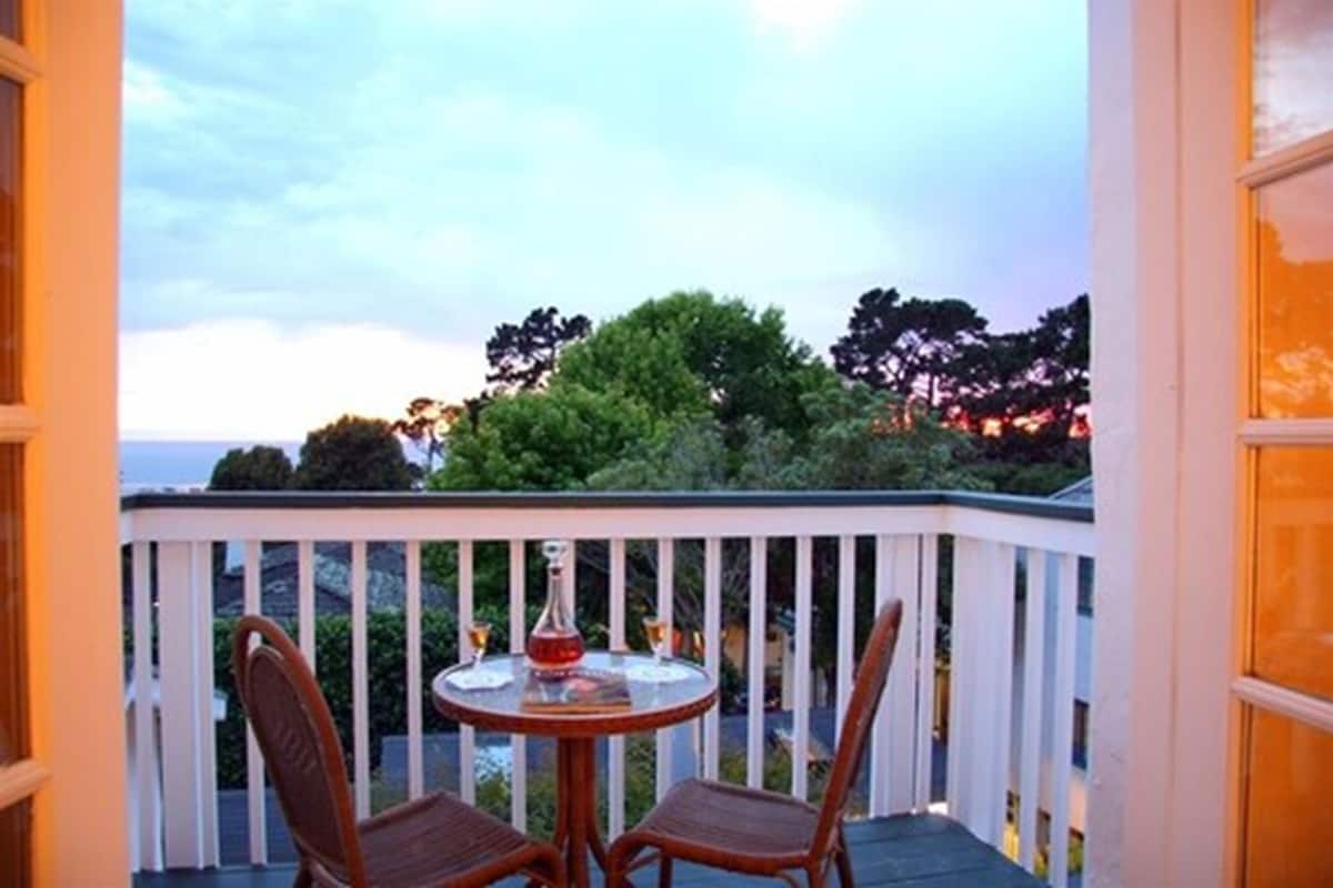 A balcony with a round table and two chairs, overlooking a scenic view of trees and the ocean.