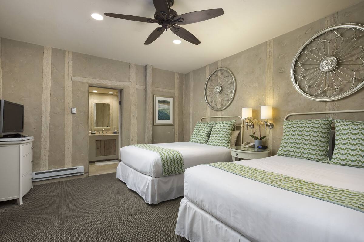 A hotel room with two beds, a ceiling fan, a television, and a bathroom visible through an open door.