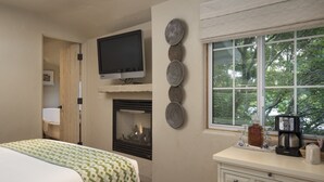 A hotel room with a bed, a fireplace, a television, decorative wall art, a window with a view of trees, and a coffee maker on the dresser. - Monte Verde Inn (Carmel)