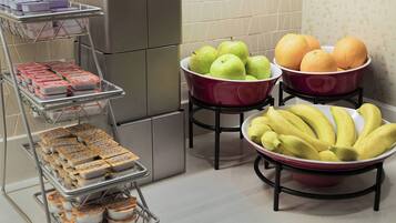 A kitchen with a refrigerator, a metal rack with packaged food, and bowls containing apples, oranges, and bananas.